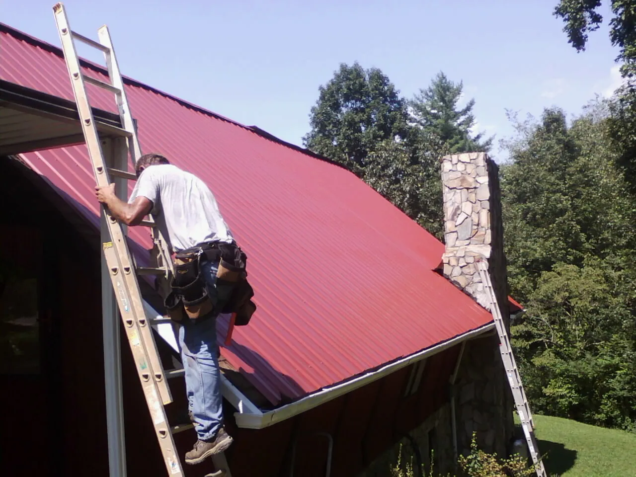Roofer on a ladder beside a red metal roof and stone chimney — Soffit & Fascia Repair by Roofing Brothers in Jarrettsville
