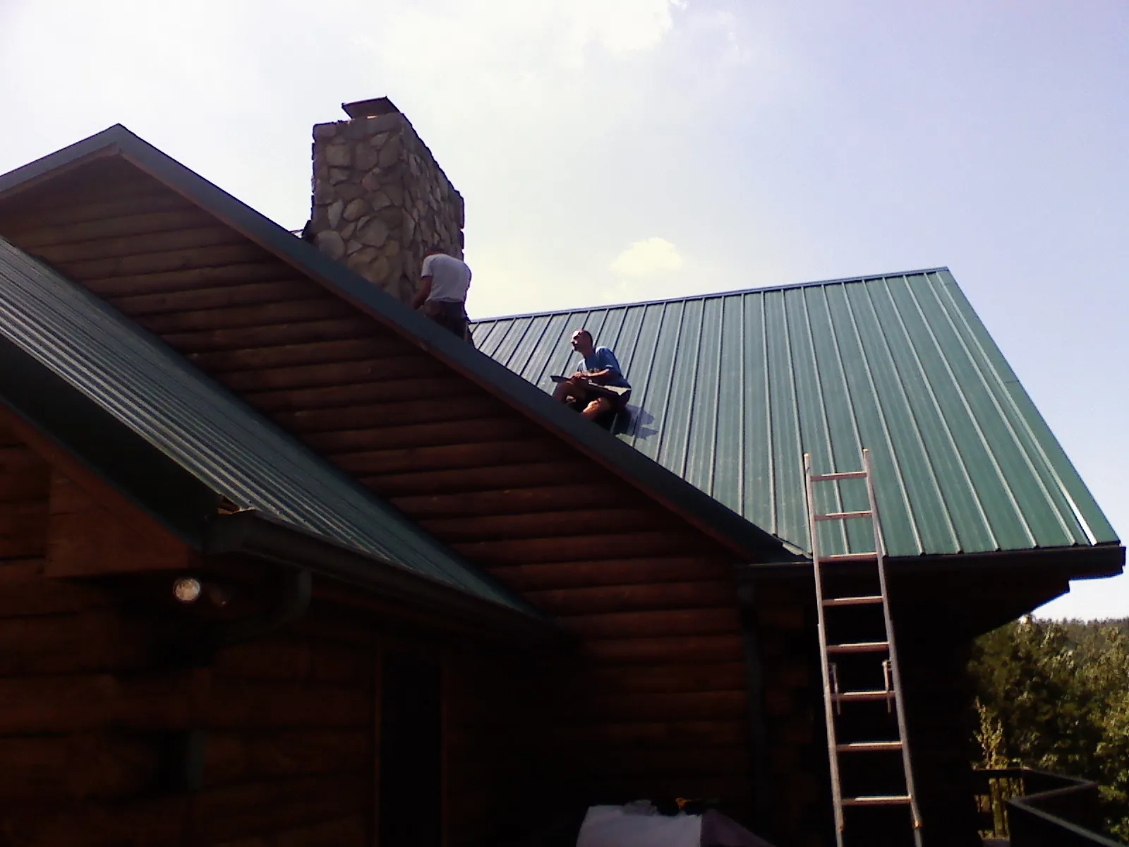 Roofers installing green metal panels beside a stone chimney on a log home in Jarrettsville